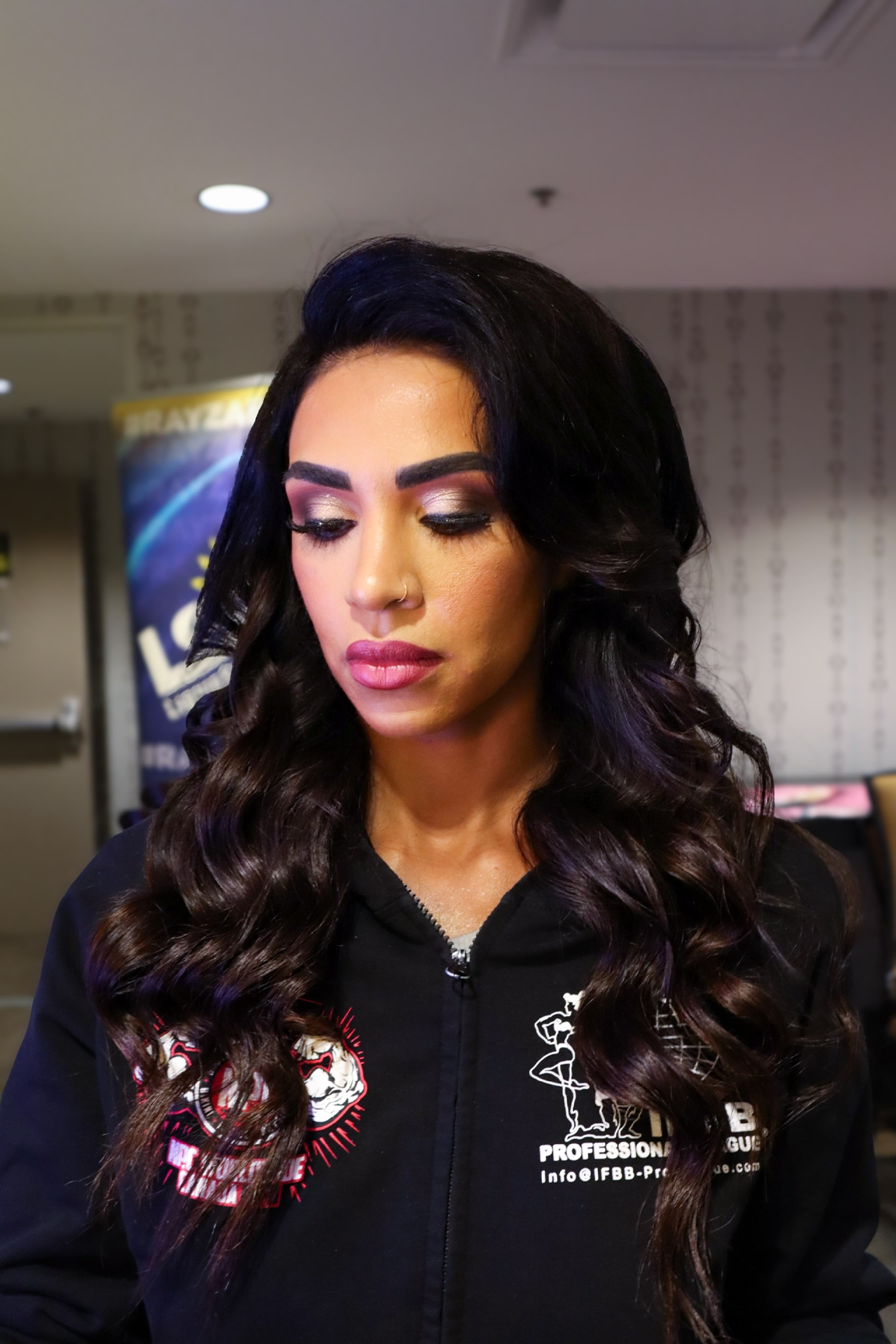 Woman with long dark wavy hair and glamorous makeup looking down in a black hoodie.