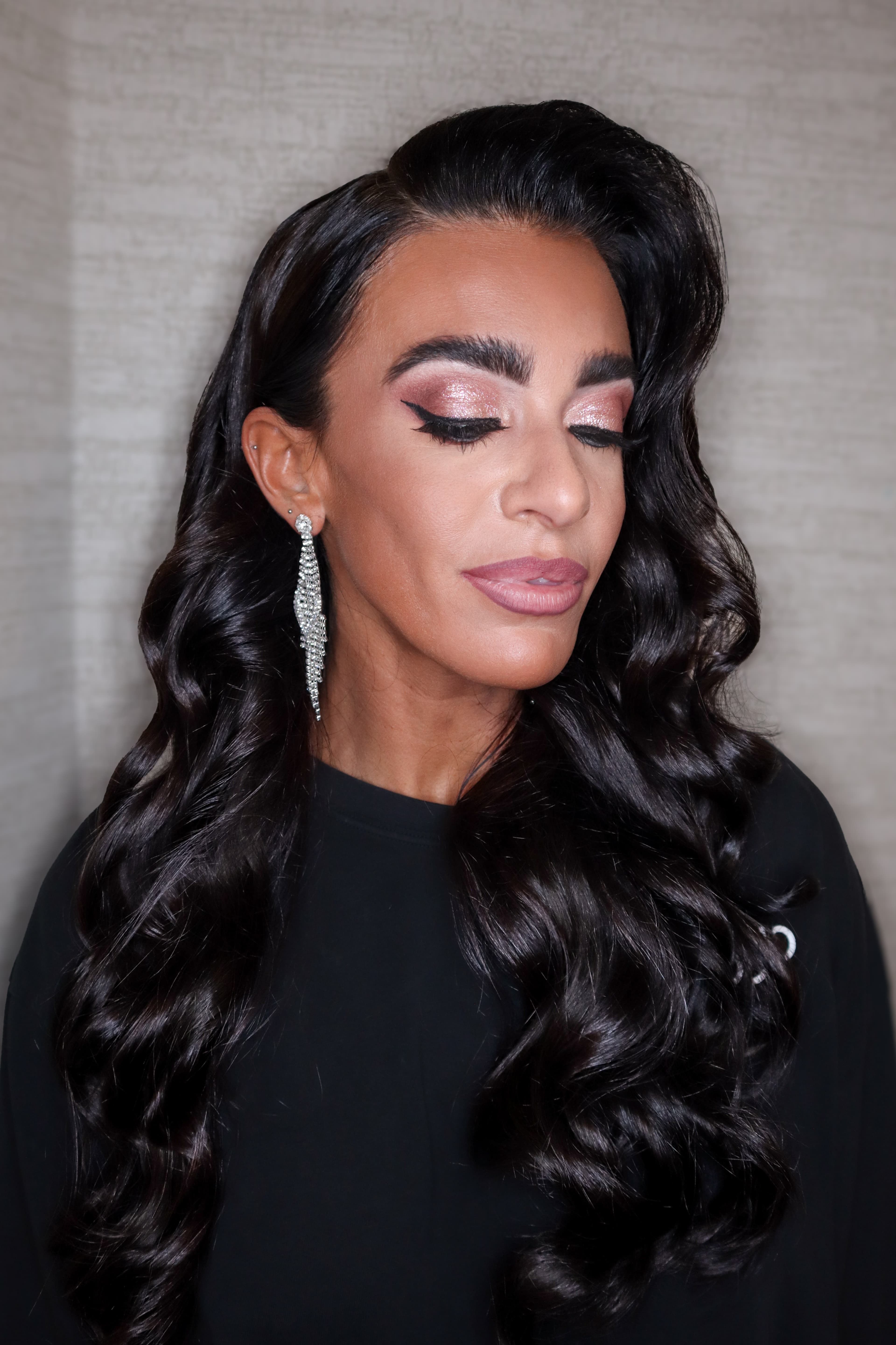 Woman with long dark waves, shimmery pink eyeshadow, winged eyeliner, and dangling silver earrings.